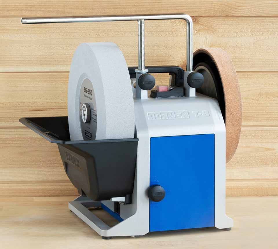 A Tormek T-8 dual-wheel stone grinder with a white abrasive wheel, a brown sanding belt, a blue front panel, and a black tray, set against a light wood background.