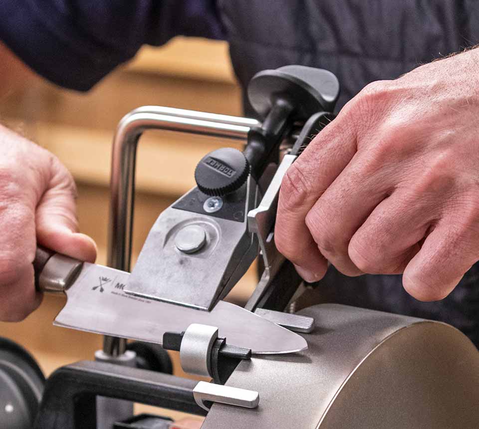 A close-up of a person sharpening a knife on a bench grinder, showing hands, the knife, and the metal sharpening tool in use.