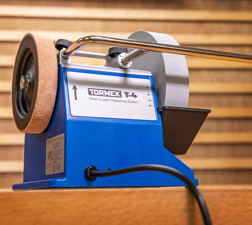 A Tormek T-4 water corded sharpening system machine with a blue metal base, a brown abrasive wheel on the left, a gray sharpening wheel on the right, and a chrome handle, shown on a wooden surface.