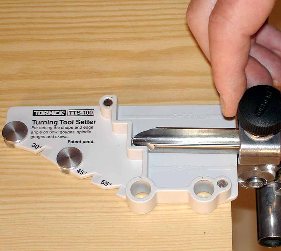 A close-up of a Tormek TTS-100 Turning Tool Setter in white and silver, being adjusted by a person's hand on a light wood surface.