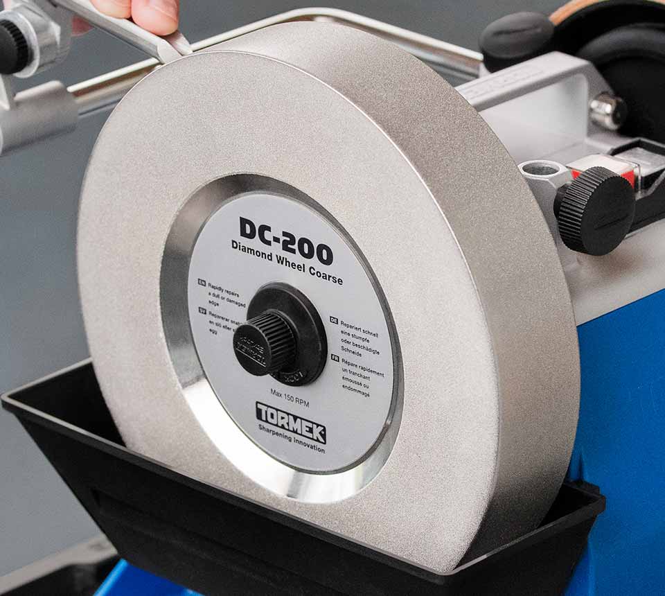 A close-up of a Tormek DC-200 diamond wheel, showing the silver abrasive wheel, black adjustment knob, and white label with product details. The wheel is mounted on a Tormek machine with a blue base and a black tray.