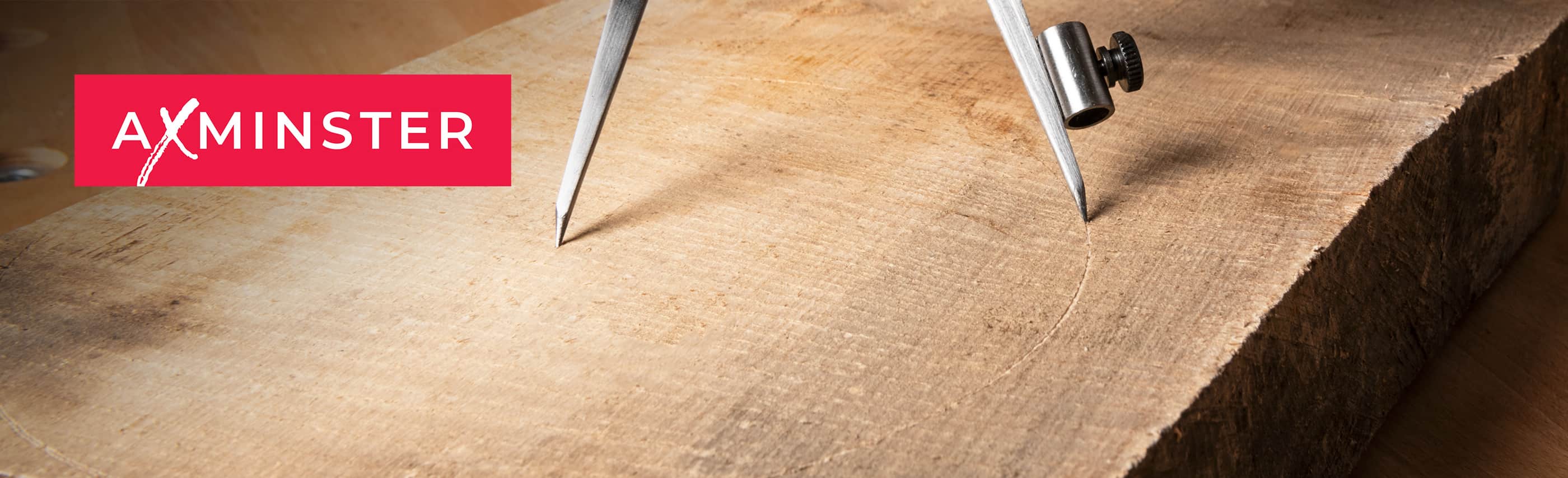 A close-up of a large natural wood slab with visible grain, showing the AXMINSTER brand logo in a red box on the left and the edges of a metal divider or compass tool at the top.