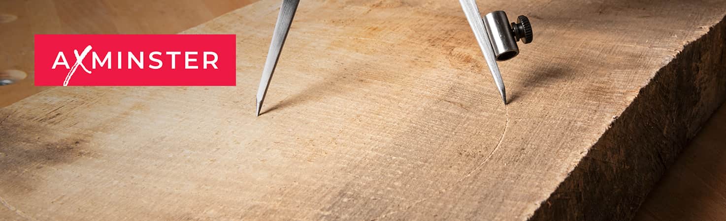 A close-up of a large natural wood slab with visible grain, showing the AXMINSTER brand logo in a red box on the left and the edges of a metal divider or compass tool at the top.