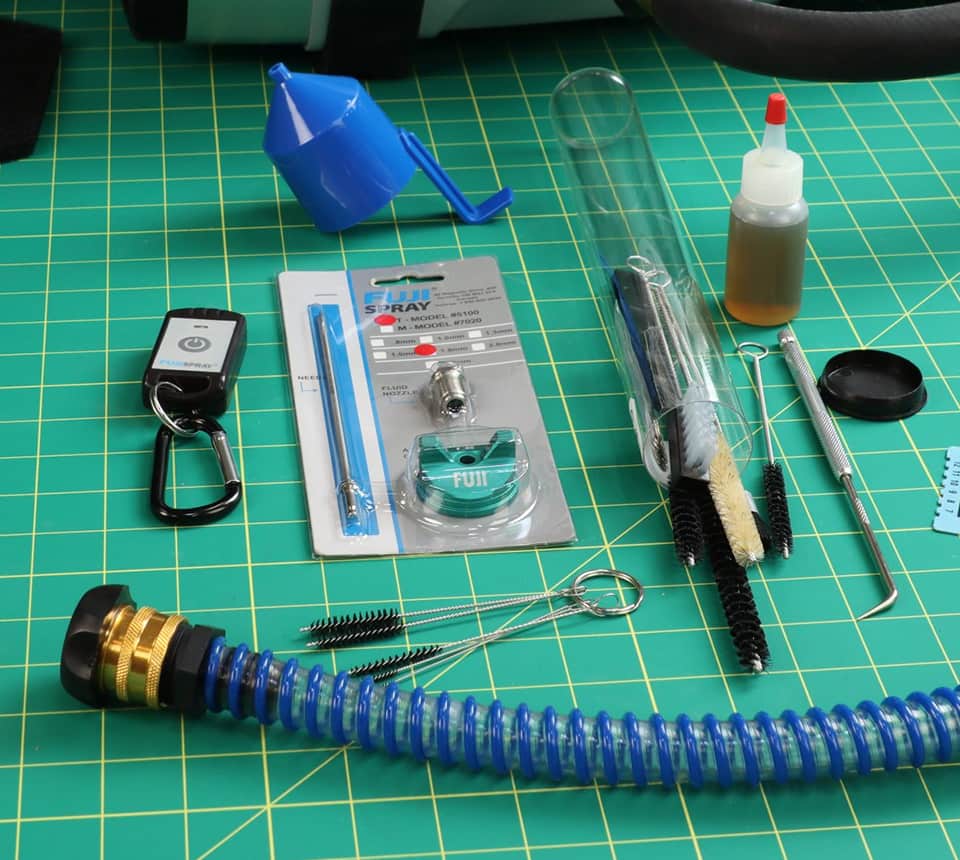 A collection of tools and parts for cleaning or repairing. A tube laid out on a green cutting mat. The set includes a blue corrugated tube, a blue funnel, a small bottle of amber liquid, various metal cleaning brushes, a black keychain, and packaged metal fittings.
