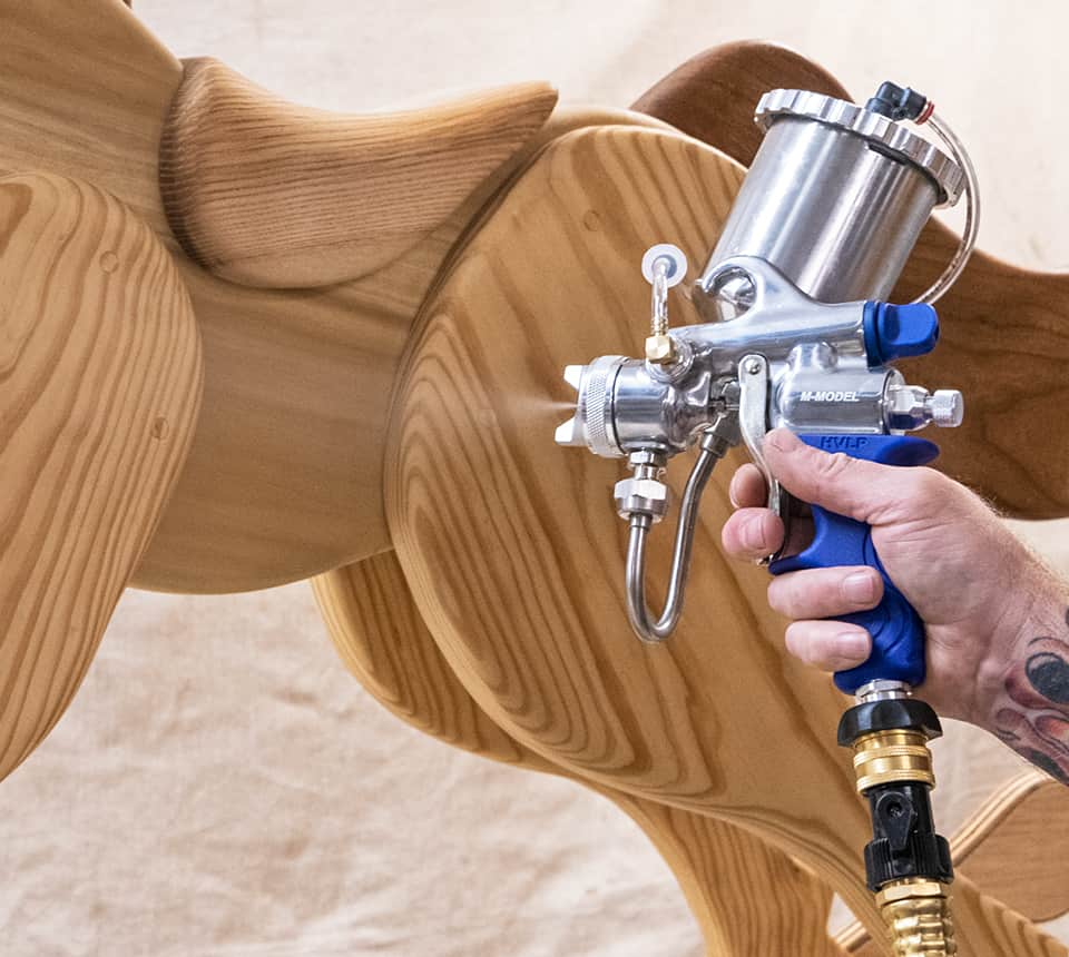 A close-up of a person's hand holding a spray gun with a blue handle and silver metal body, applying finish to wooden rocking horse. The spray gun has a clear acrylic hopper, silver metal spray nozzle, and a brass hose connector.