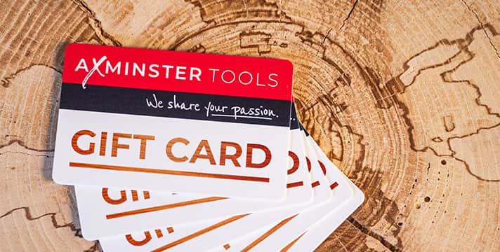 A stack of AXMINSTER TOOLS gift cards fanned out on a rustic wood slice background. The top card is red, black, and white with the AXMINSTER TOOLS logo and the words 'We share your passion.