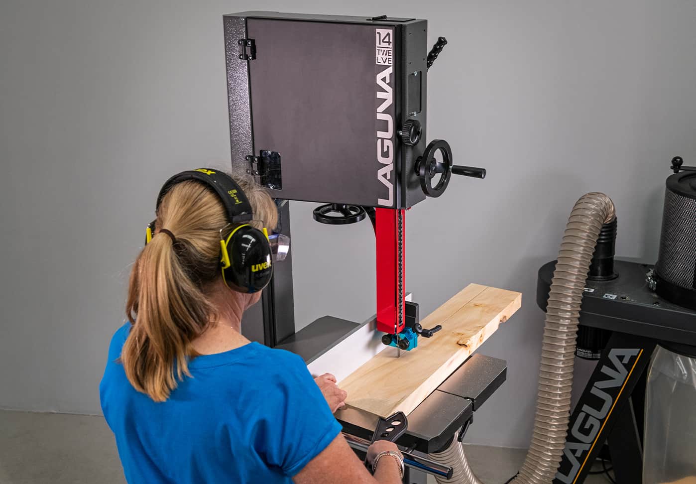 A woman wearing protective earmuffs operates a Laguna 150 band saw cutting a piece of wood, with a dust collection system visible on the right against a gray studio background.