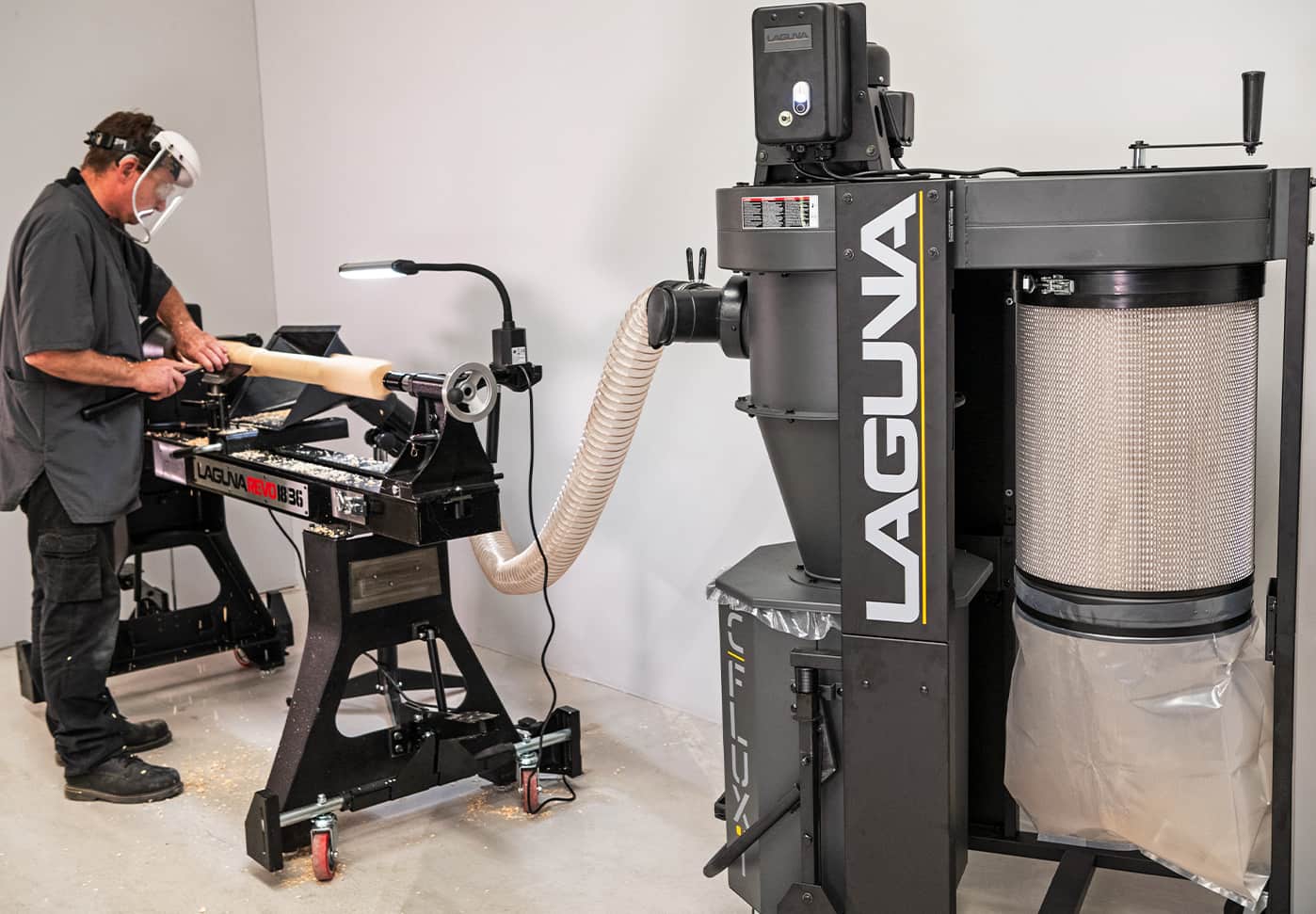 A woodworker wearing a face shield operates a lathe, with wood shavings being drawn into a large black Laguna dust collector in a workshop.