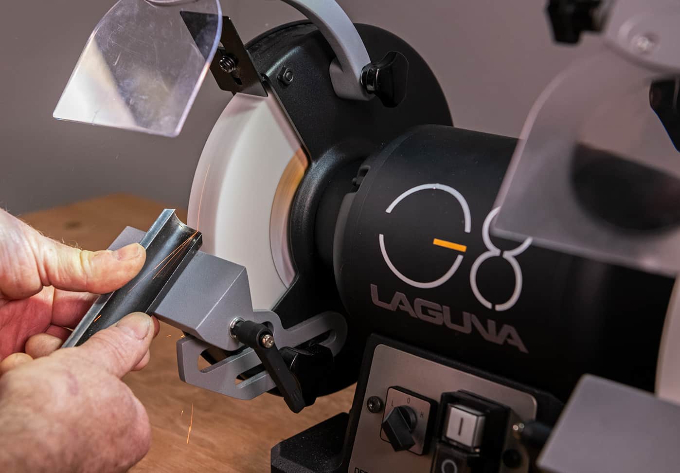 A close-up of a person using a Laguna CG8 bench grinder to sharpen a chisel, with the Laguna brand name clearly visible on the black machine body.