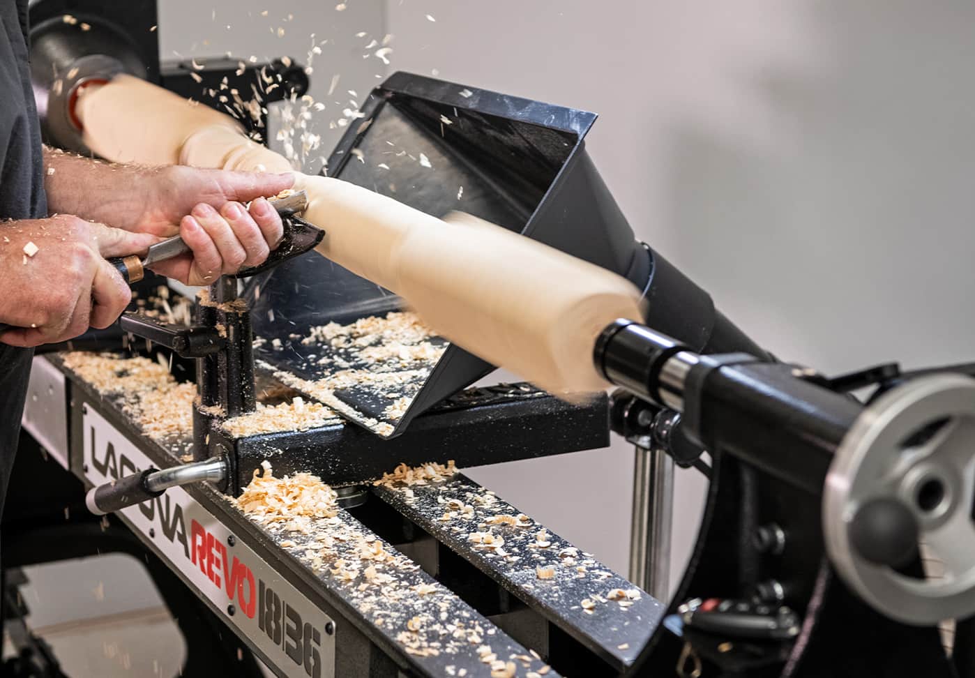 A close-up action shot of a woodturning lathe in operation, showing a person's hands guiding a piece of wood as it spins and shavings fly off. The brand 'LAPAVA REVO 1836' is clearly visible on the machine's bed.