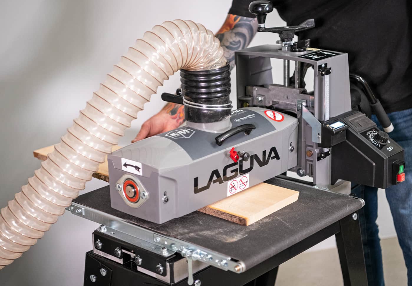 A Laguna wood planer with a flexible beige dust collector hose, shown in use with a person operating it in a workshop setting.