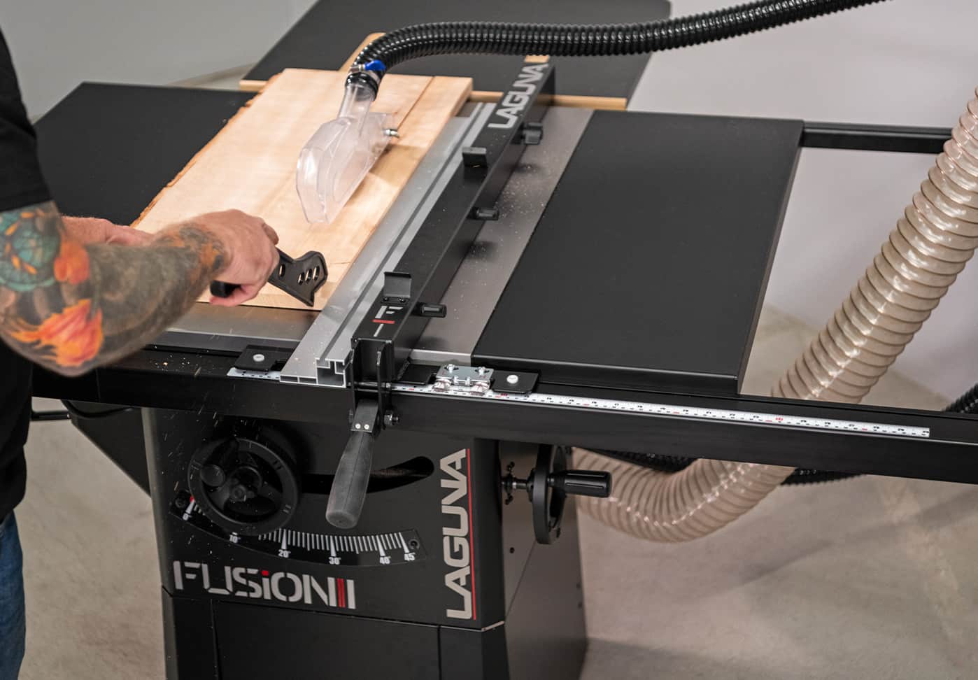 A person guides a wood sheet through a Laguna Fusion table saw with a clear guide and attached dust collection hose in a workshop setting.