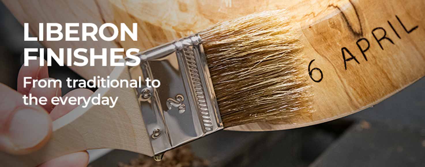 A close-up of a hand holding a wooden paintbrush with a silver metal ferrule. The brush has '6 APRIL' printed on the wooden handle, and the background is dark with white text reading 'LIBERON FINISHES' and 'From traditional to the everyday'.