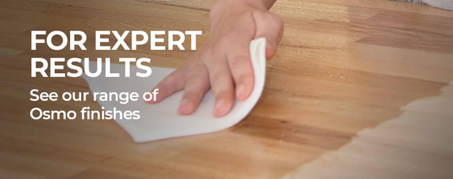 A hand using a white cloth to wipe a light brown wooden surface, with text on the left reading 'FOR EXPERT RESULTS See our range of Osmo finishes'.