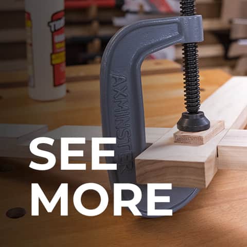 A close-up of a grey clamp holding light wood blocks on a workbench, with a blurred workshop background and large white text reading 'SEE MORE' overlaid across the lower portion.