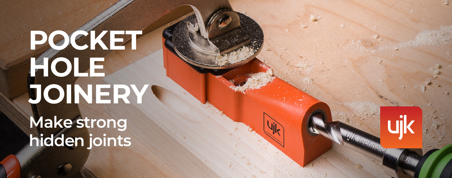 A close-up action shot of a woodworking tool in use, showing a pocket hole joinery bit creating a joint in a piece of wood. The bright orange jig holds the drill bit, with wood shavings scattered on the workbench.