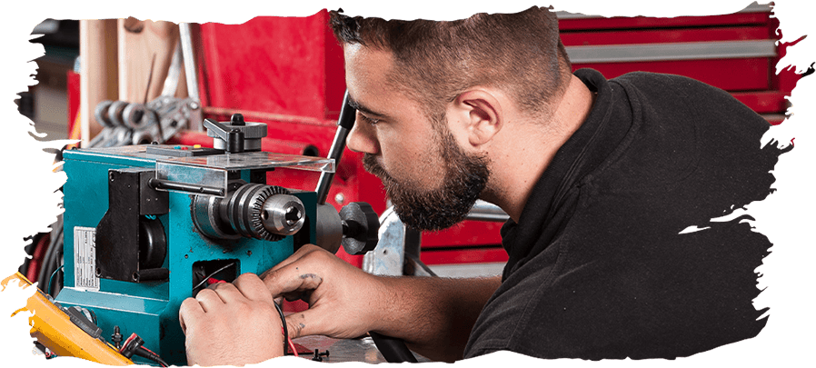 A bearded man in a black shirt works closely with a teal and silver metal lathe machine in a workshop setting with a red tool chest in the background.