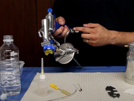 A person's hands holding and pointing to a silver metal spray gun attachment with blue and yellow accents, set against a dark background with a workbench containing a water bottle, a small bottle, and other tools.