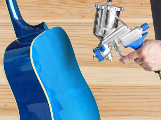 A close-up of a hand holding a silver and blue spray gun and applying paint to the body of a blue acoustic guitar against a light wood background.