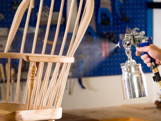 A hand holding a spray gun with a metal paint can, spraying a light natural wood spindle-back chair in a workshop setting with a blue pegboard background.