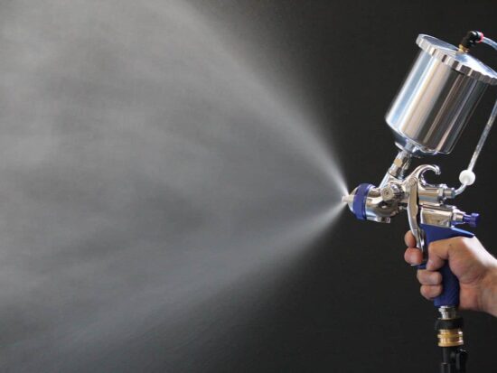 A close-up of a hand holding a silver and blue spray gun, spraying a fine mist of liquid against a dark grey background.