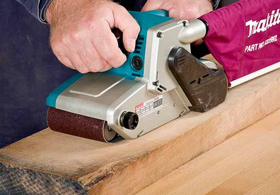 A person using a Makita belt sander to smooth a piece of wood. The sander has a teal and silver body with a brown sanding belt, and a pink Makita dust collection bag is attached to the side.