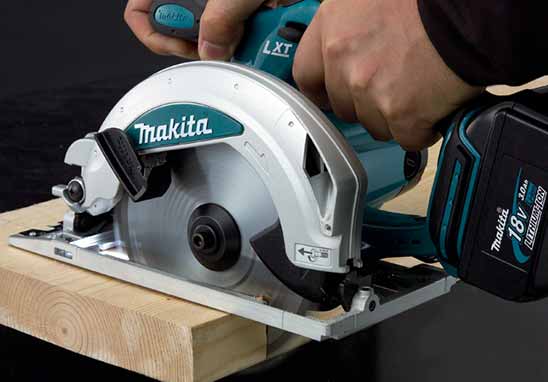 A person using a Makita circular saw with a wood guide rail on a piece of wood, showing the teal and silver Makita tool in action.