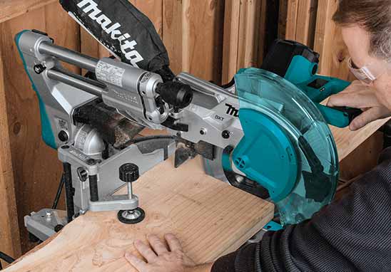 A Makita circular saw miter saw in teal and silver cutting a piece of wood, with a person's hands and head visible guiding the wood in a workshop setting.