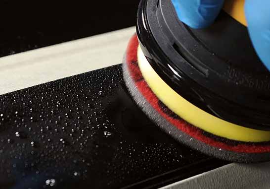 A close-up action shot of a black circular buffer pad with a yellow and red foam edge, being used on a glossy black surface by a hand wearing a blue glove.