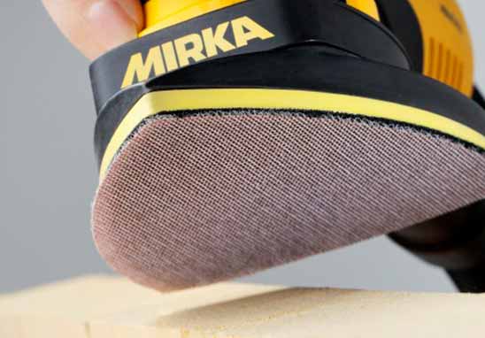 A close-up of a yellow and black Mirka sander with a brown abrasive sanding disc, being used to sand a light-colored wooden surface, with a hand holding the tool.