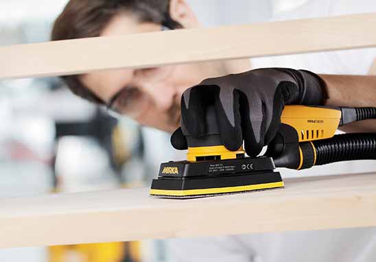 A person wearing a black work glove uses a yellow and black Mirka sander on a light wood board in a workshop setting.