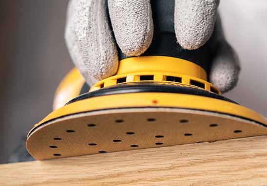 A close-up of a gloved hand using a yellow and black orbital sander on a light wood surface, showing the sanding dust collection port and sanding pad.