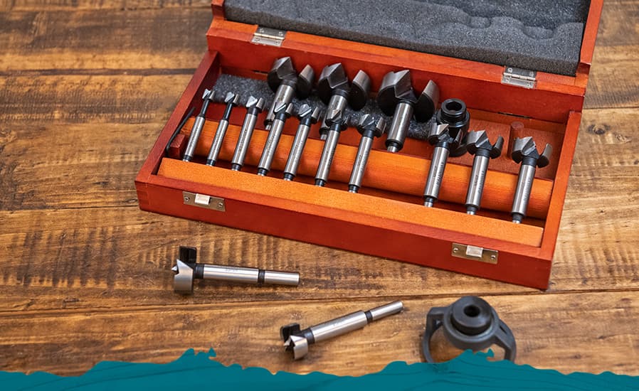 A set of metal router bits displayed in an open reddish-brown wooden presentation case on a wooden tabletop, with a few bits placed outside the case.