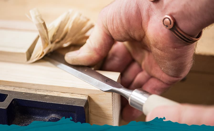 A close-up of a person's hand using a metal chisel to carve or shape a piece of light wood, with wood shavings visible and a dark workbench in the background.