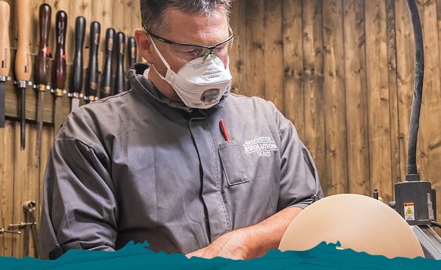 A man wearing a grey work shirt and a white dust mask operates a woodturning lathe, with a wooden wall of hand tools behind him.